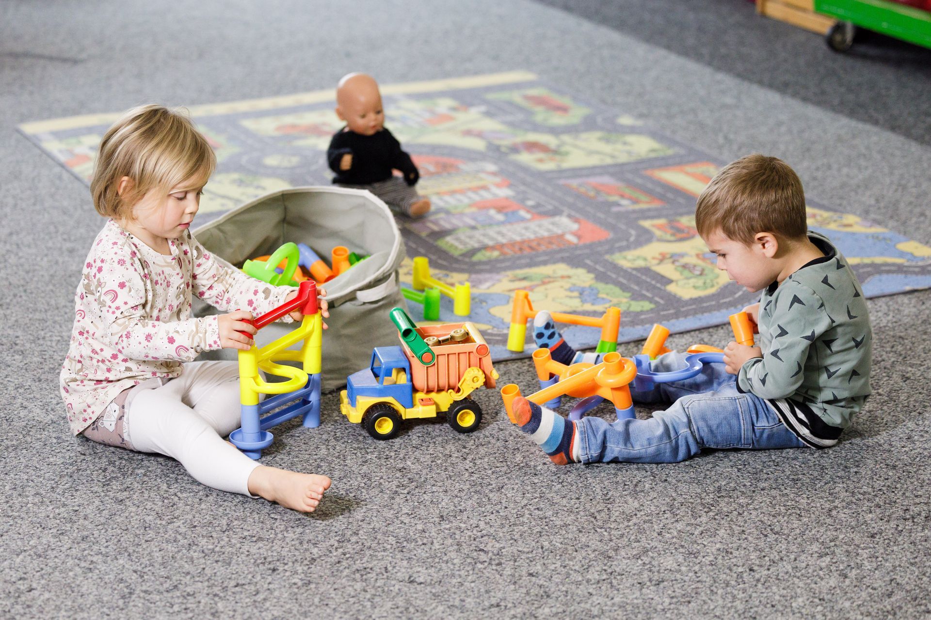 Zwei kleine Kinder spielen mit buntem Spielzeug auf einem Teppich in einem Spielbereich.