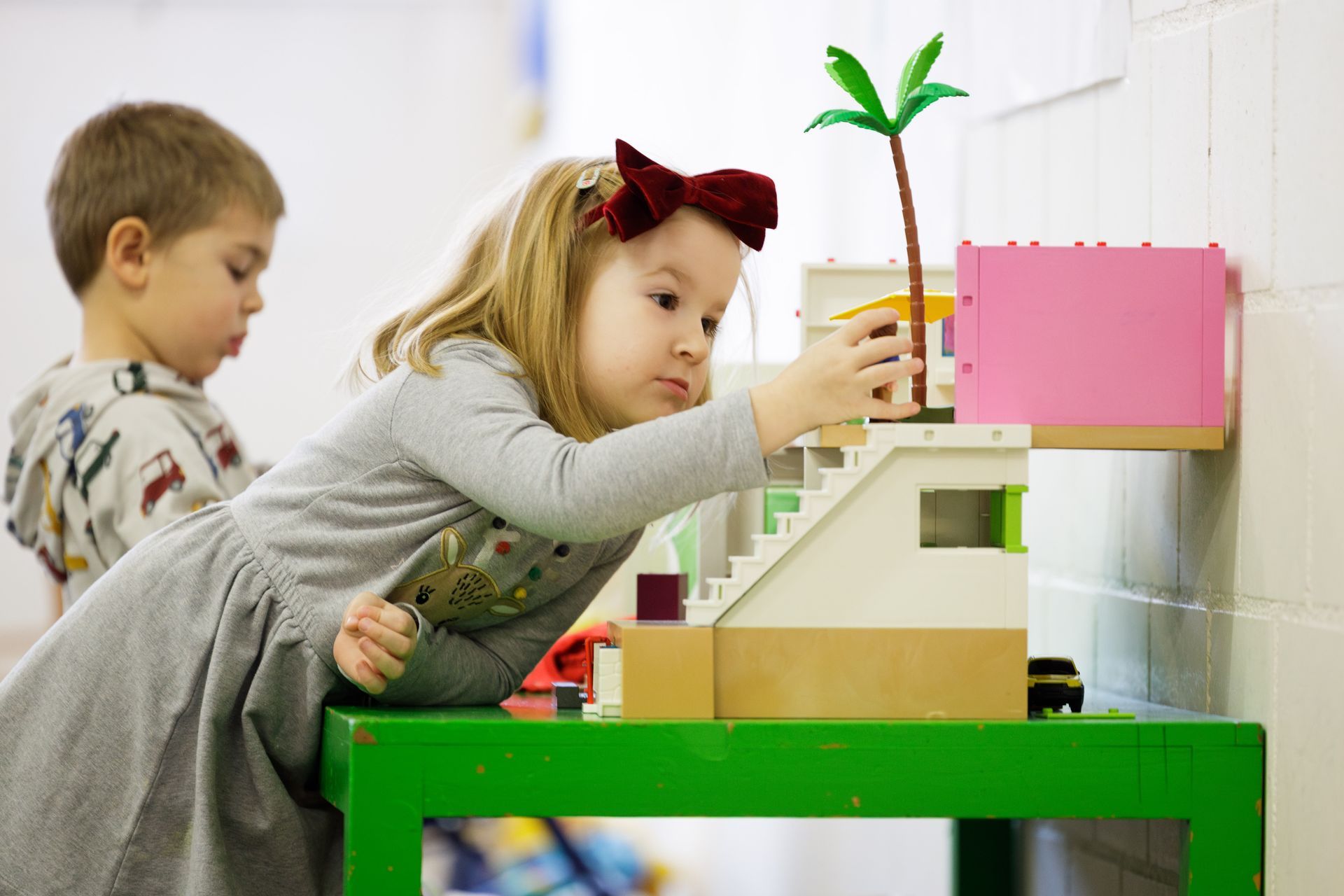 Zwei Kinder spielen mit Bauklötzen an einem grünen Tisch im Innenbereich. Ein Kind stellt eine Palme auf das Gebäude.