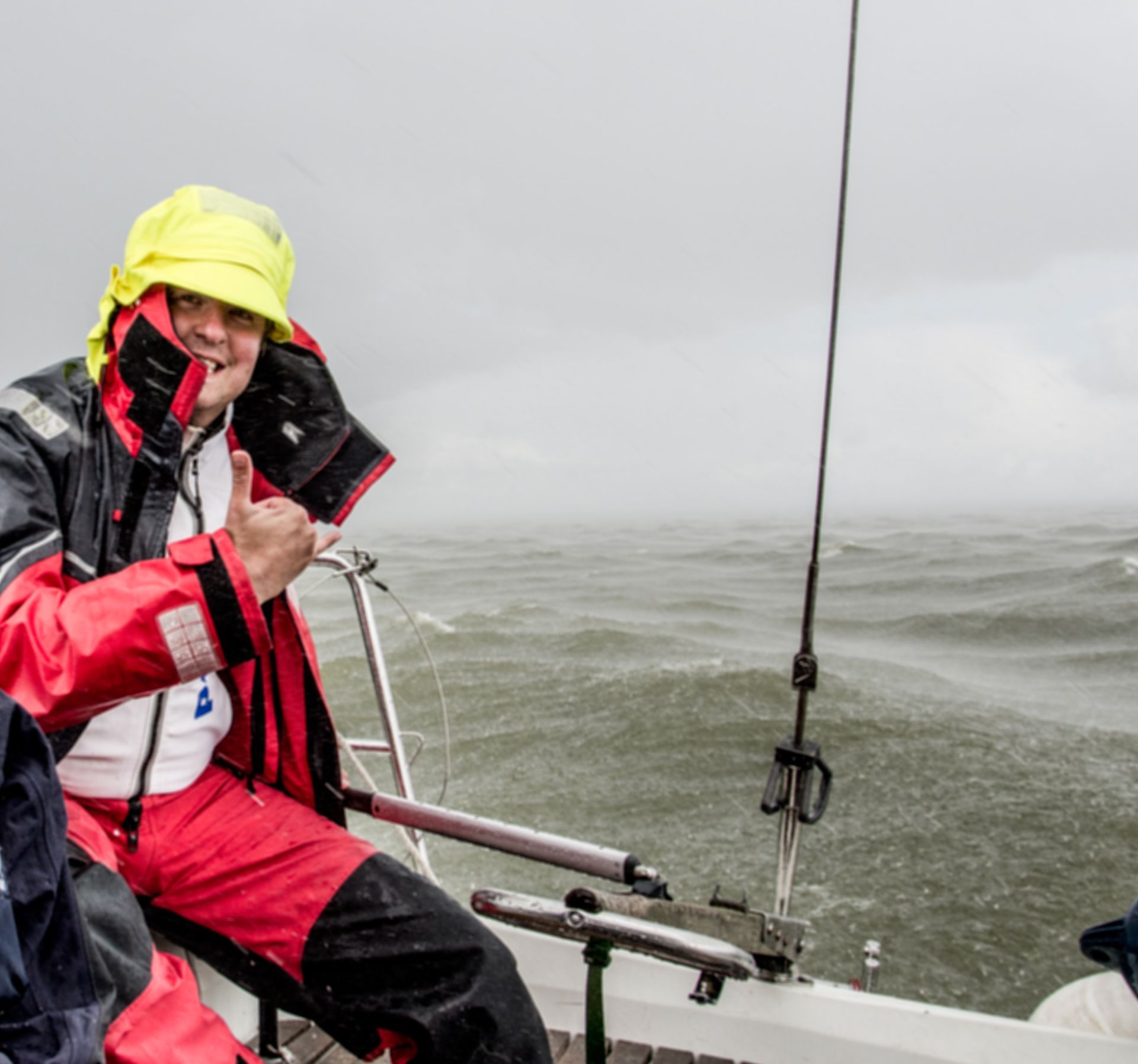 Een persoon op een zeilboot in een storm steekt zijn duim omhoog; hij draagt rode en gele waterdichte kleding.