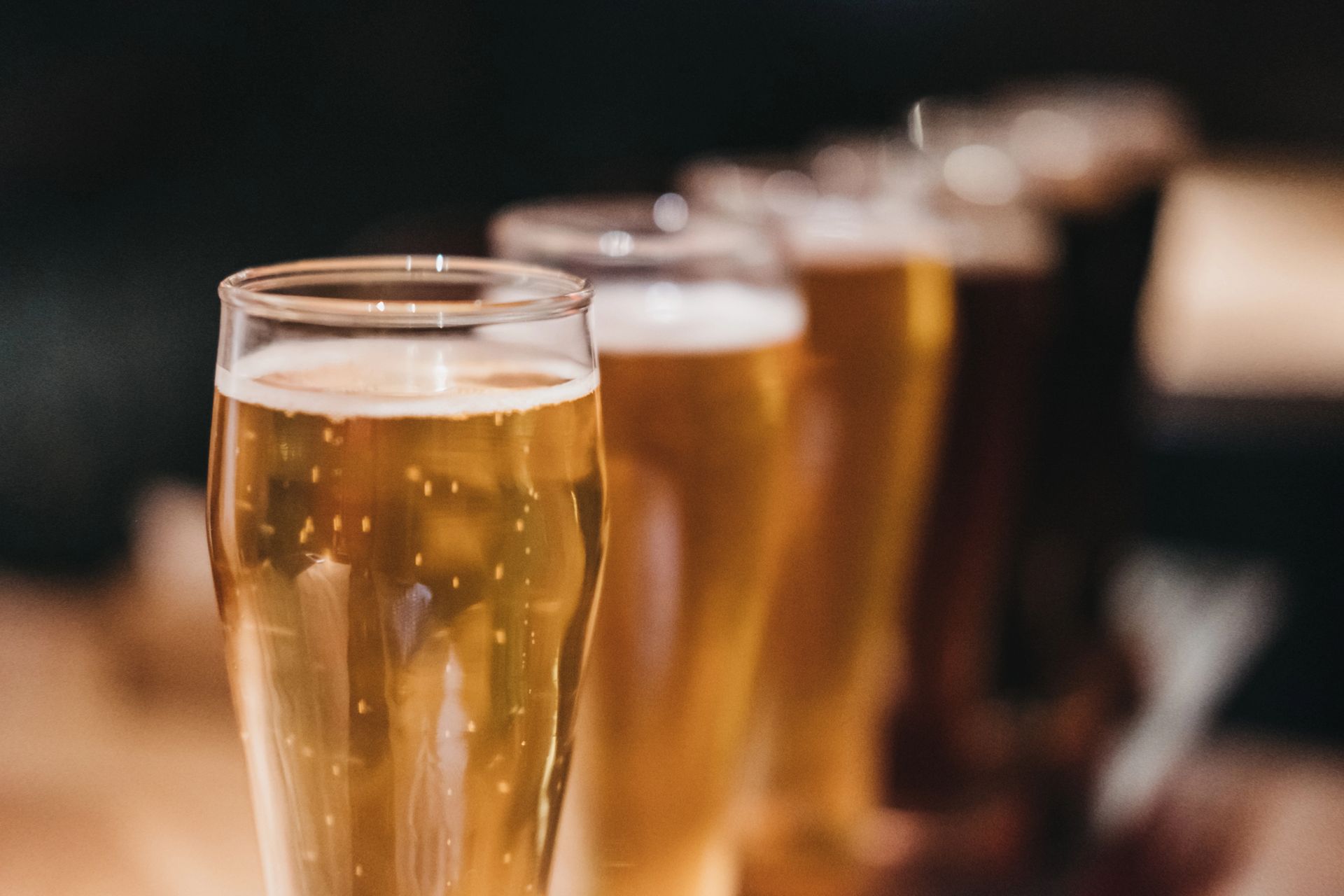 File di bicchieri da birra dorati allineati sul bancone di un bar, in una luce soffusa e con lo sfondo sfocato.