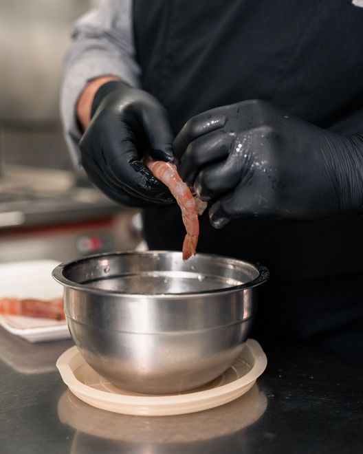 Mani guantate che maneggiano cibo sopra una ciotola di metallo in un ambiente di cucina.