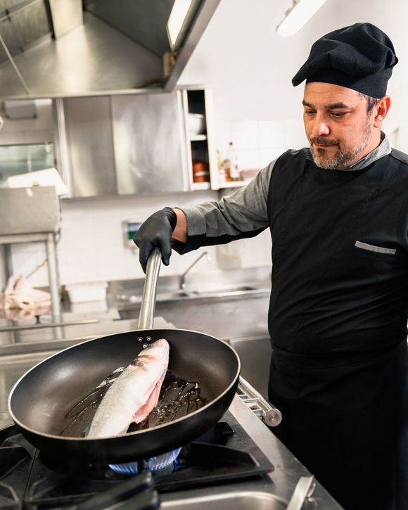 Lo chef cucina un filetto di pesce in padella su un fornello a gas in una cucina in acciaio inossidabile.