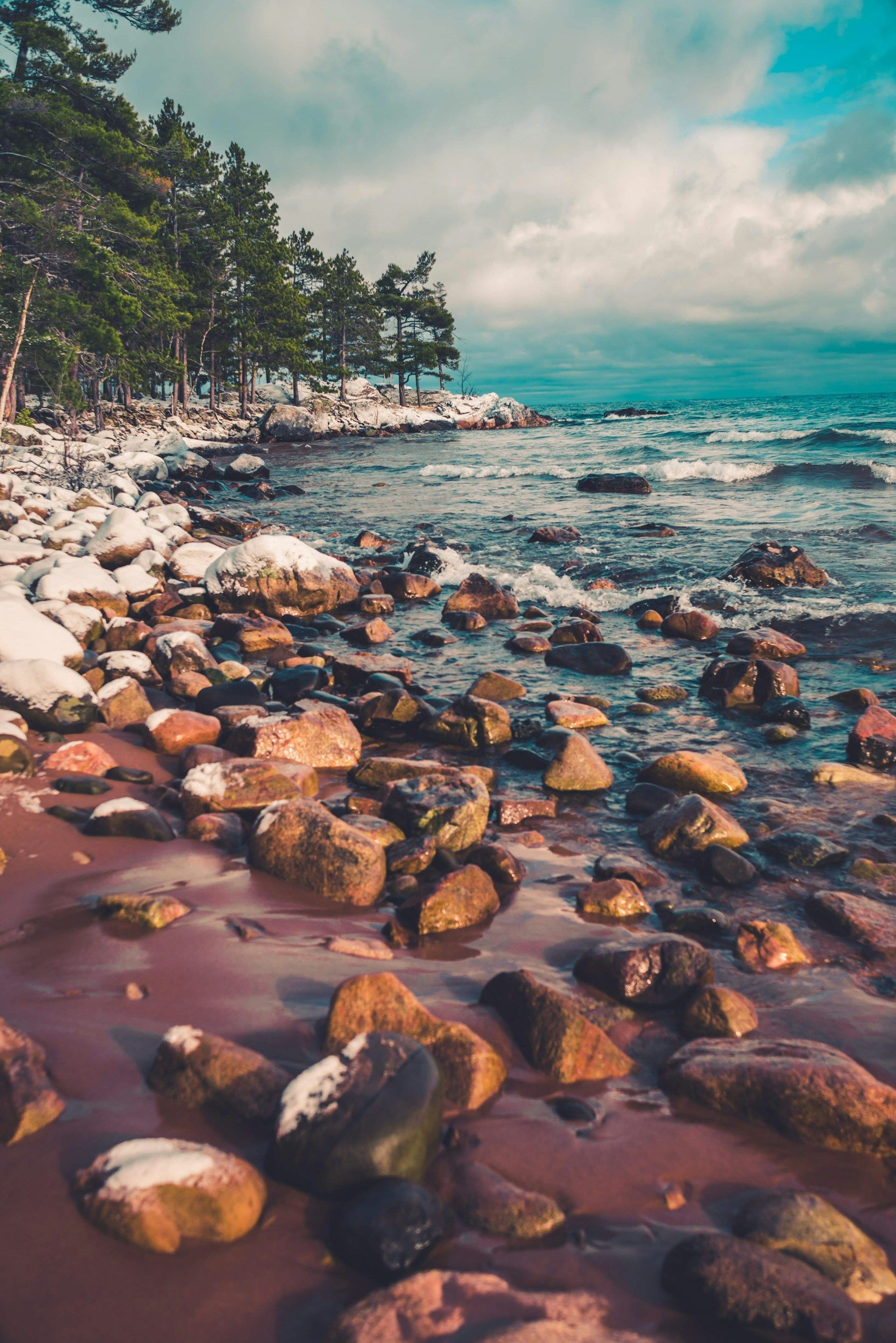 Rocky shoreline with a beach in the foreground and a line of evergreen trees along the coast under a cloudy blue sky.