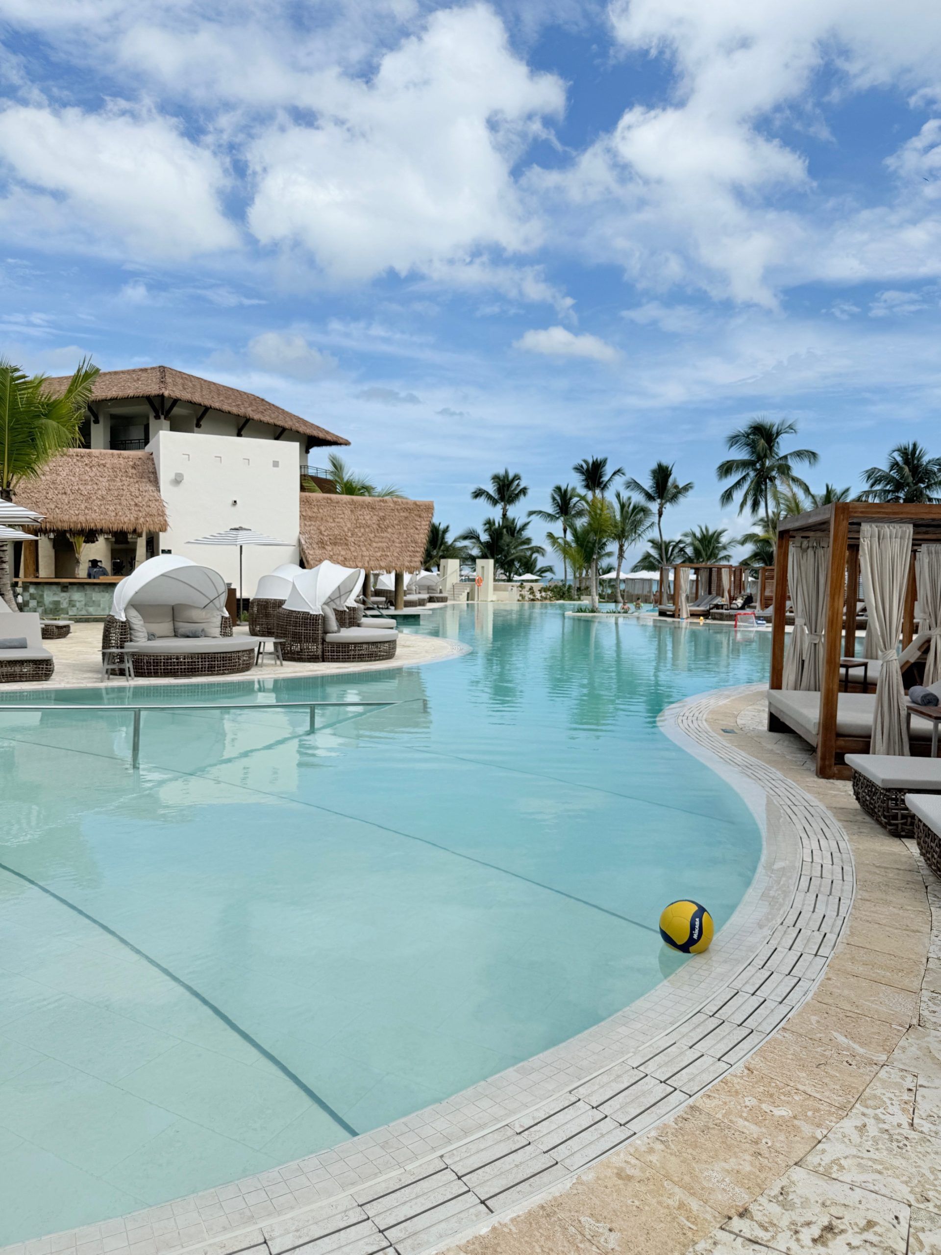 Resort pool with clear blue water, lounge chairs, cabanas, and palm trees under a partly cloudy sky