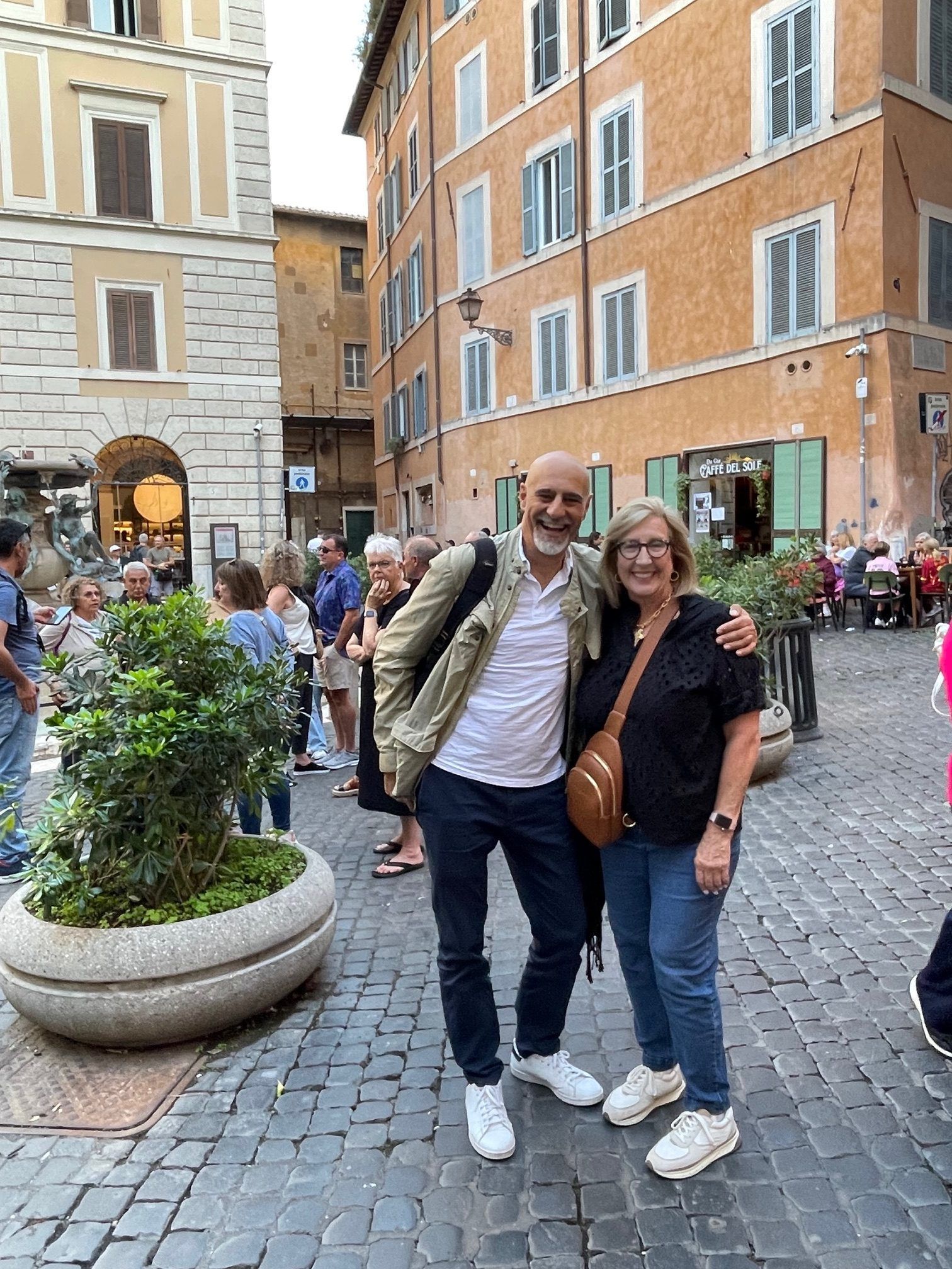 Two smiling people posing on a cobblestone street in front of warm-colored buildings and a small plaza fountain