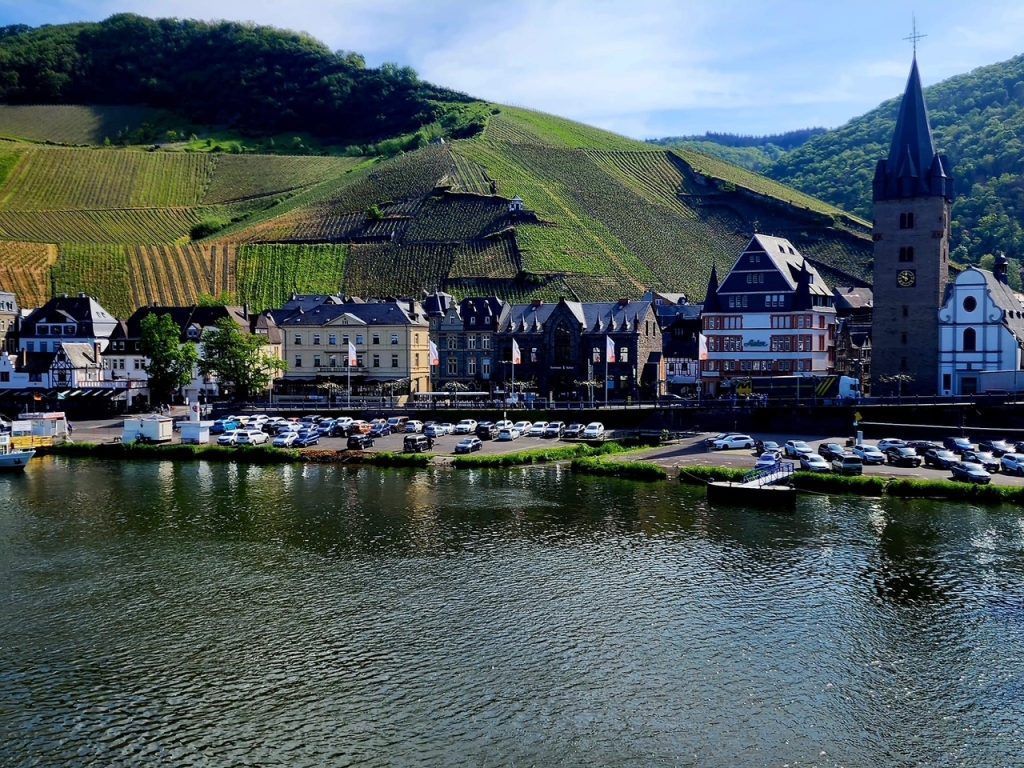 Riverside village with white buildings and a church tower beside calm water and green hills