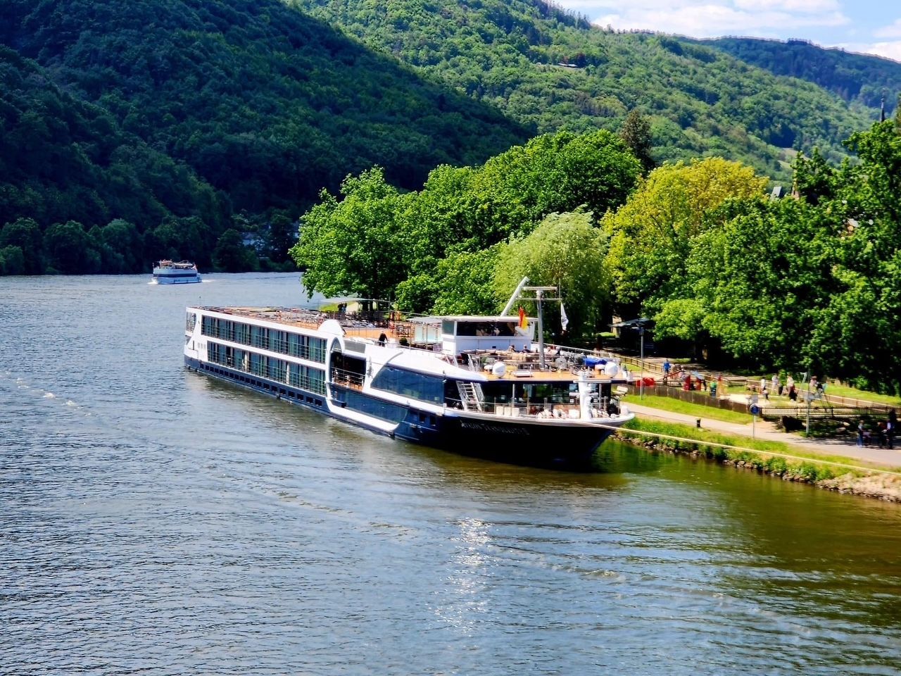 Long riverboat docked by a green riverbank with forested hills in the background