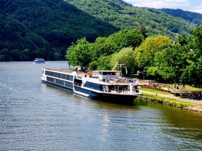 River cruise boat docked by a green forested riverbank and mountains