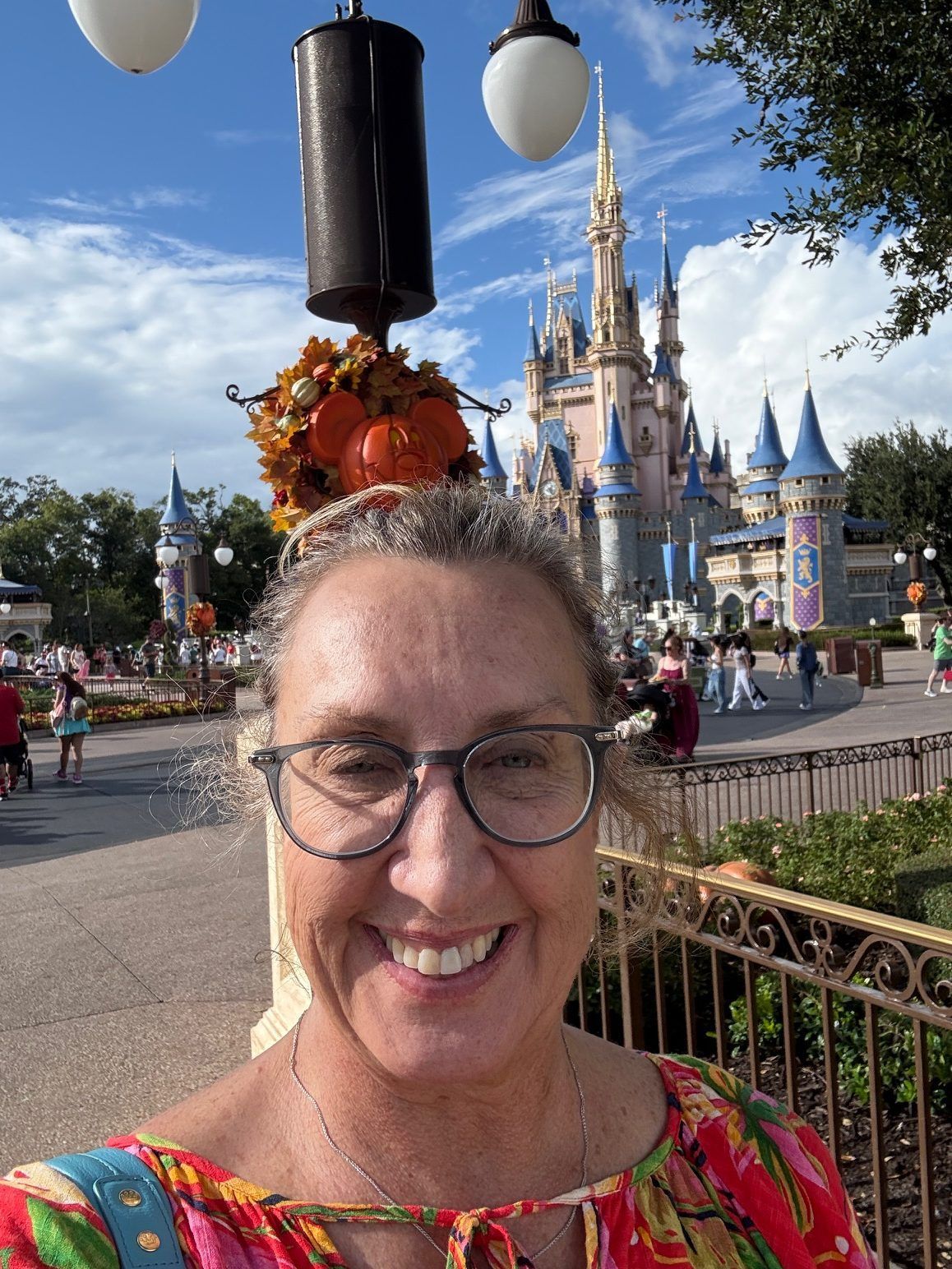Smiling woman in glasses at a Disney castle, wearing a red floral top and festive headband.