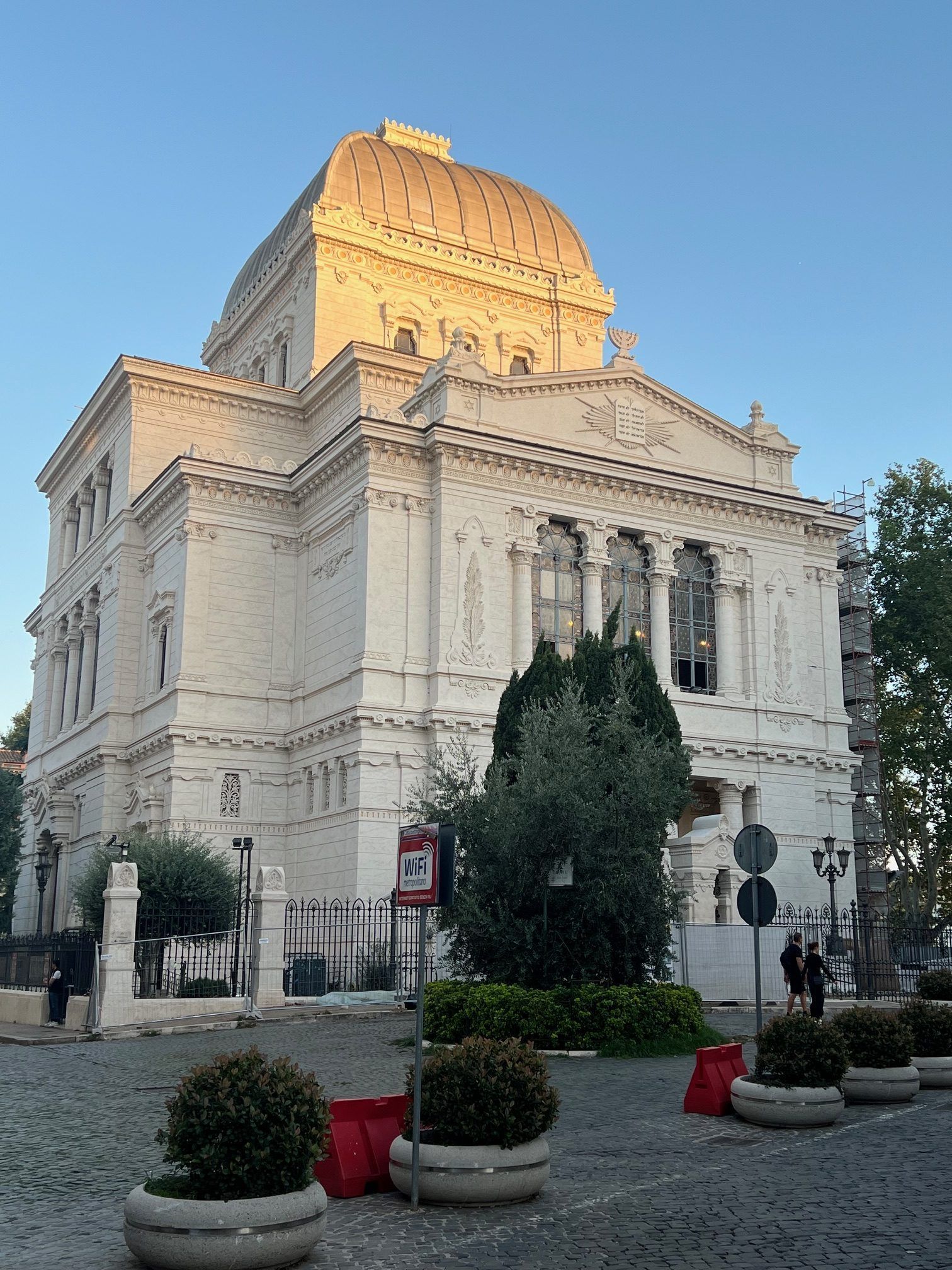 White historic building with ornate dome and columns, seen at sunset behind planters and trees.