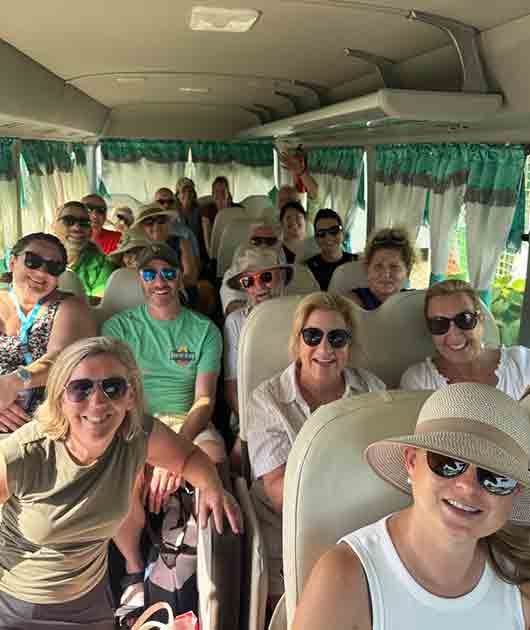 Group of smiling people seated on a bus, many wearing sunglasses and light-colored shirts.