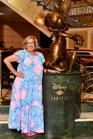 Woman in a blue floral dress posing beside a bronze Disney statue on a cruise ship lobby floor