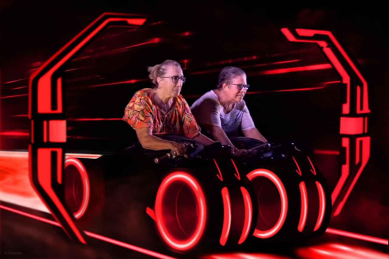 Two people seated in a glowing red futuristic ride or vehicle, with neon outlines and a dark background