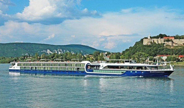 Long blue-and-white passenger ferry on a river with green hills and a hillside town in the background