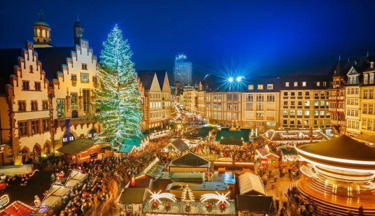 Festive Christmas market at night with a lit tree, crowded stalls, and glowing historic buildings