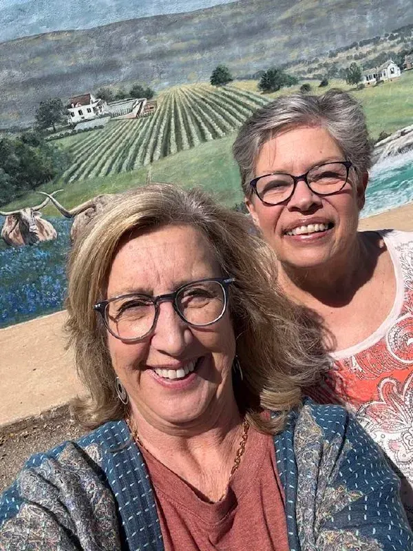 Two smiling women in glasses posing before a vineyard mural on a sunny day