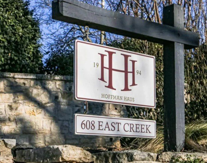 Hoffman Haus sign on stone wall with “608 East Creek” beneath, framed by trees and sky