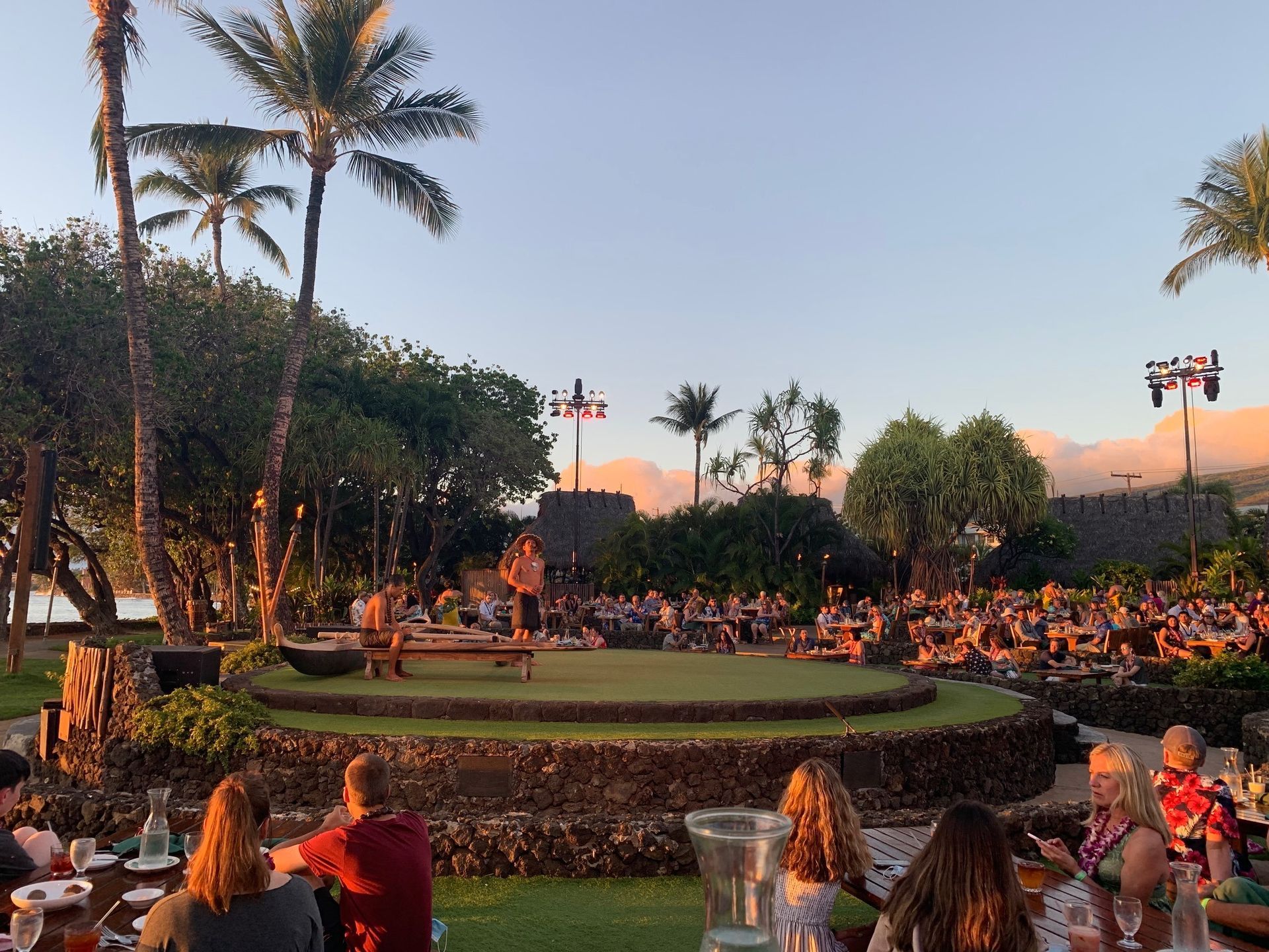 Outdoor luau stage at sunset with palm trees and a large seated audience