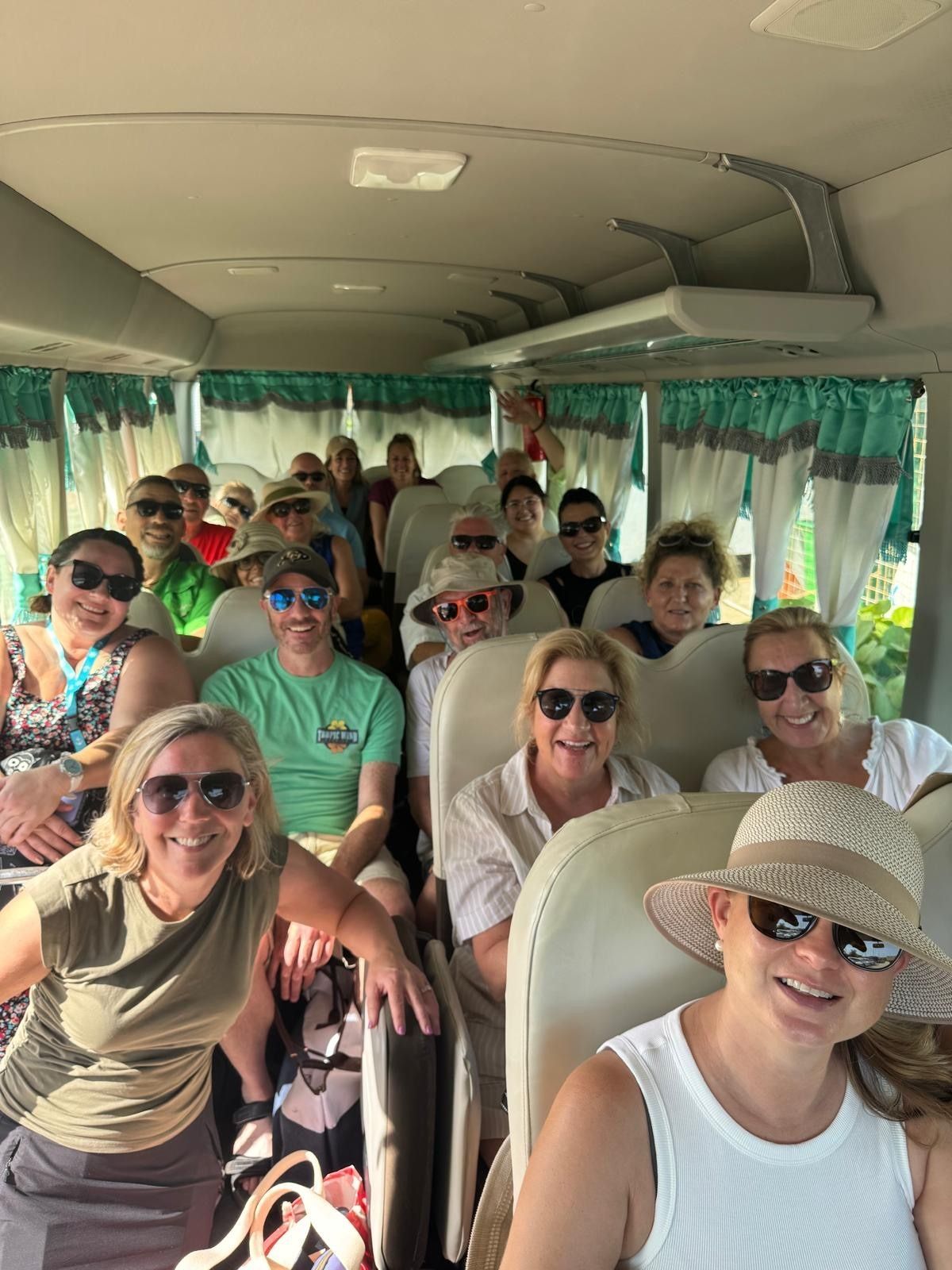 A group of smiling tourists seated inside a small bus with green curtains.
