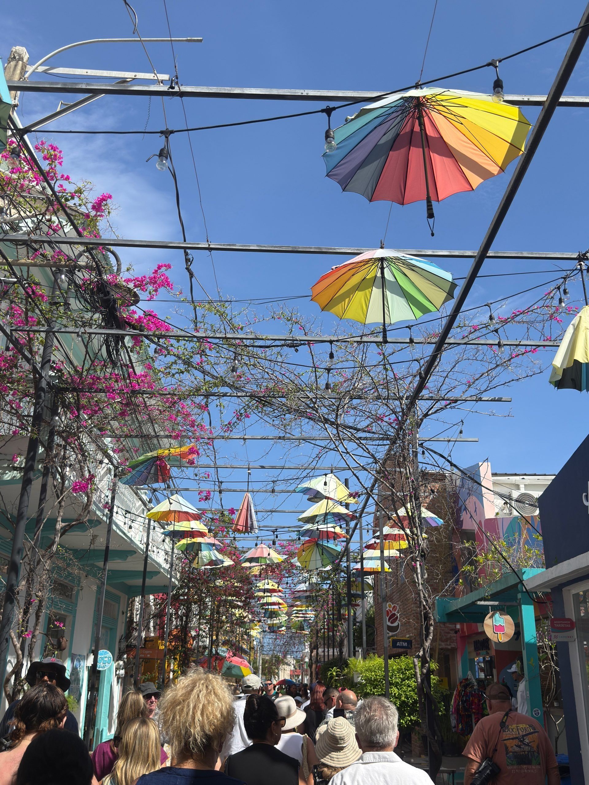 Colorful umbrellas hang over a busy outdoor market street under a bright blue sky.