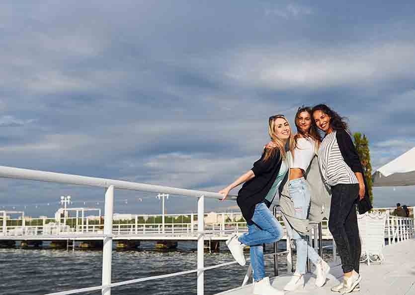 Three friends posing on a waterfront bridge, smiling with city skyline in the background.