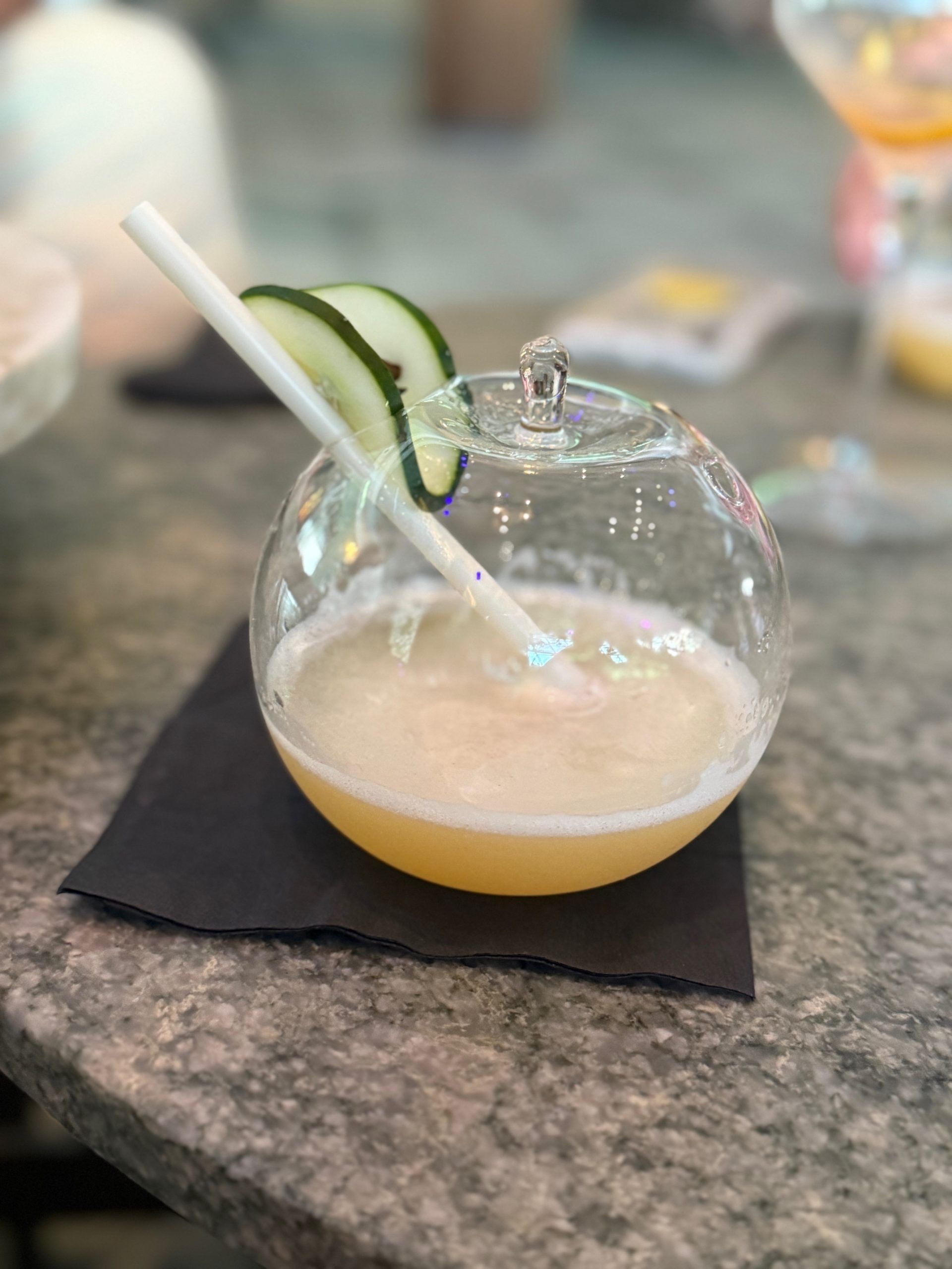 Round glass cocktail with cucumber garnish and straw on a black napkin on a stone table.