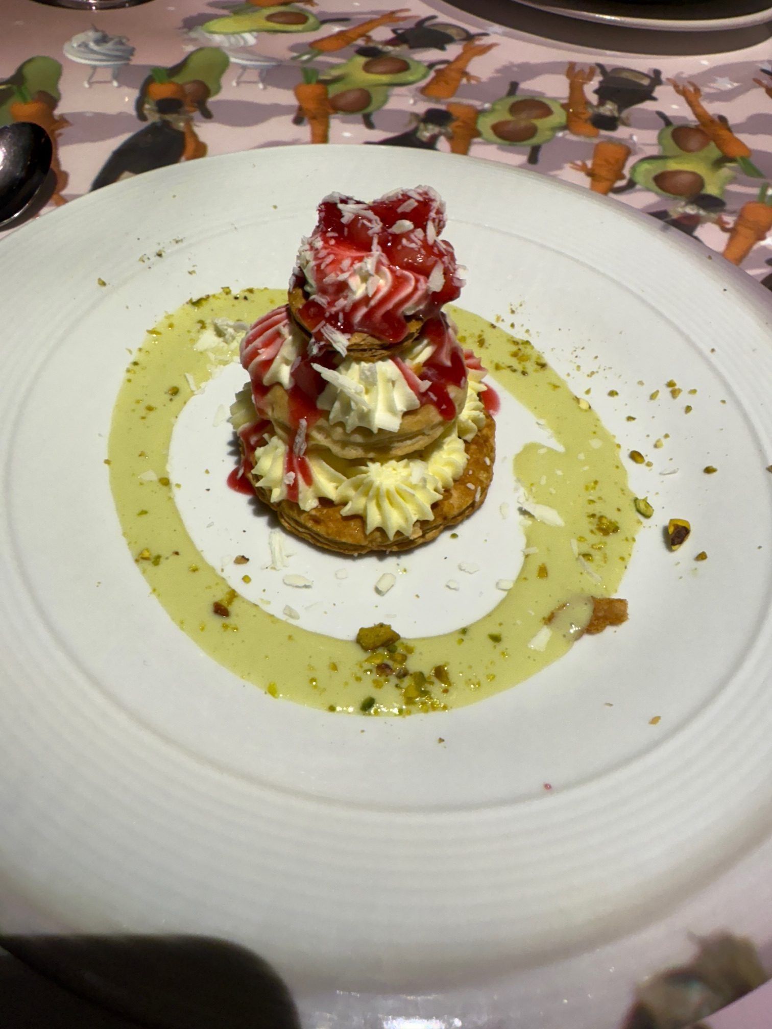 Dessert with cream and strawberry sauce on a white plate, drizzled with green sauce and pistachio crumbs.