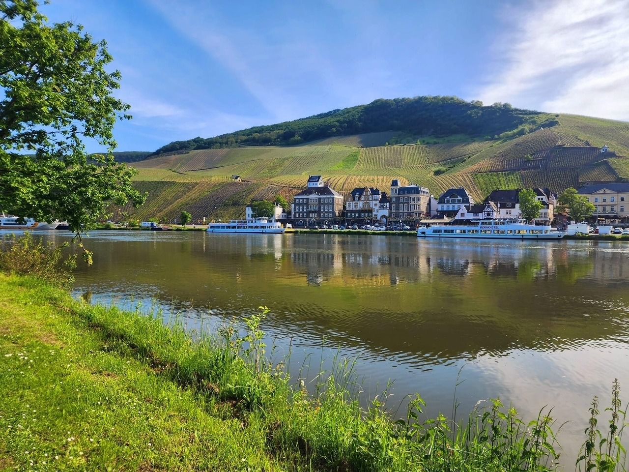Riverside village with colorful houses, a hill behind it, and trees reflected in calm water under a blue sky