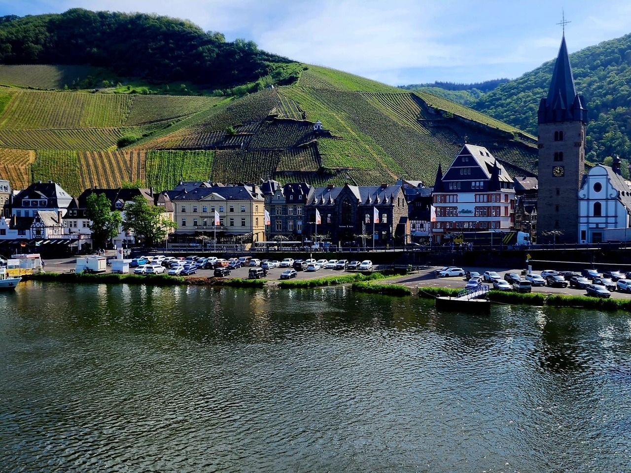 Riverside village with boats, white houses, and a church tower below green vineyard-covered hills