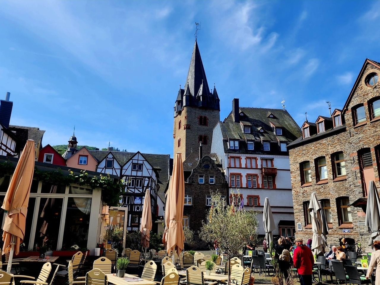 Historic town square with half-timbered buildings, a tall church spire, and café seating under a blue sky
