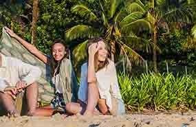 Two people sitting on a sandy beach with palm trees and lush greenery behind them.