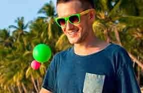 Smiling man in green sunglasses and blue shirt outdoors with balloons and palm trees