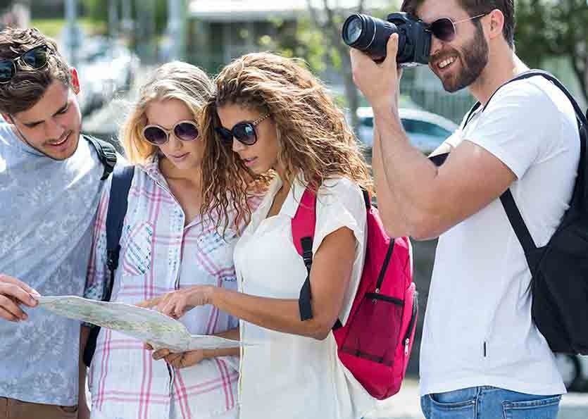 Three tourists study a map while a man with a camera takes their photo outdoors.