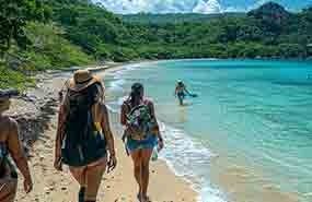 People walking along a sandy tropical beach beside clear turquoise water and green hills