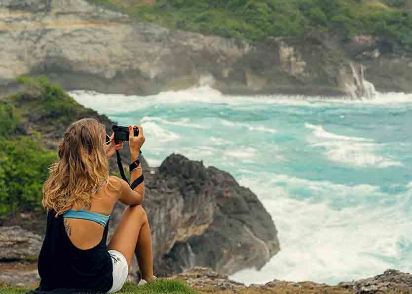 Person with curly hair photographing a turquoise ocean from a rocky cliff