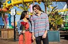 Two people standing and smiling at each other in a colorful amusement park with yellow roller coaster tracks