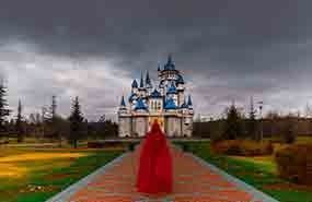 Red-cloaked figure on a walkway leading to a blue-and-white castle under a dark sky