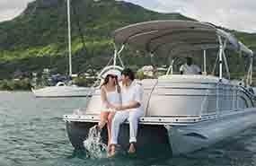 Couple sitting on the front of a white pontoon boat on calm water near a green shoreline