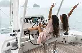 Two people in a bright white yacht cabin, sitting at the helm and raising their arms as the boat moves.
