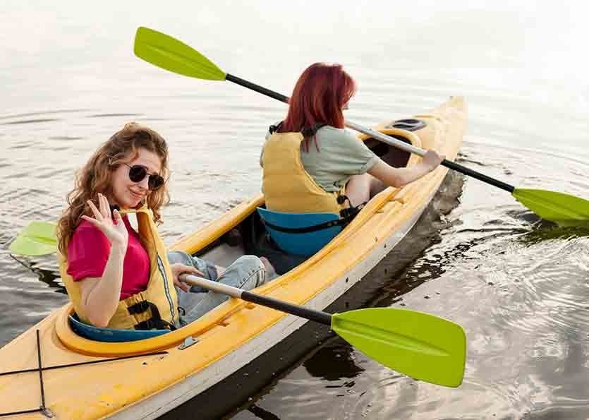 Two people in a yellow kayak on calm water, paddling with green oars; one looks back and smiles.
