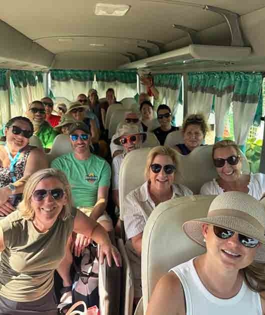 Smiling group seated on a tour bus, wearing sunglasses and hats, with green curtains and bright daylight.