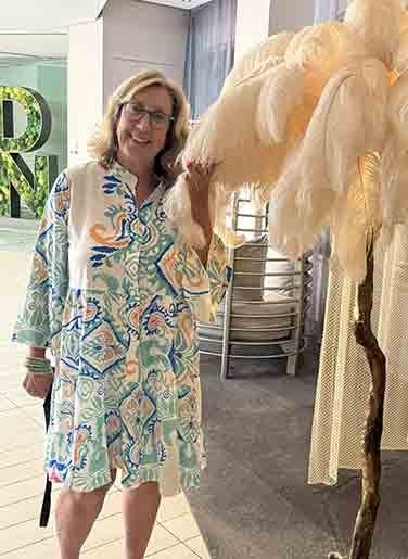 Woman in a colorful patterned dress standing beside a large feathery plant sculpture indoors