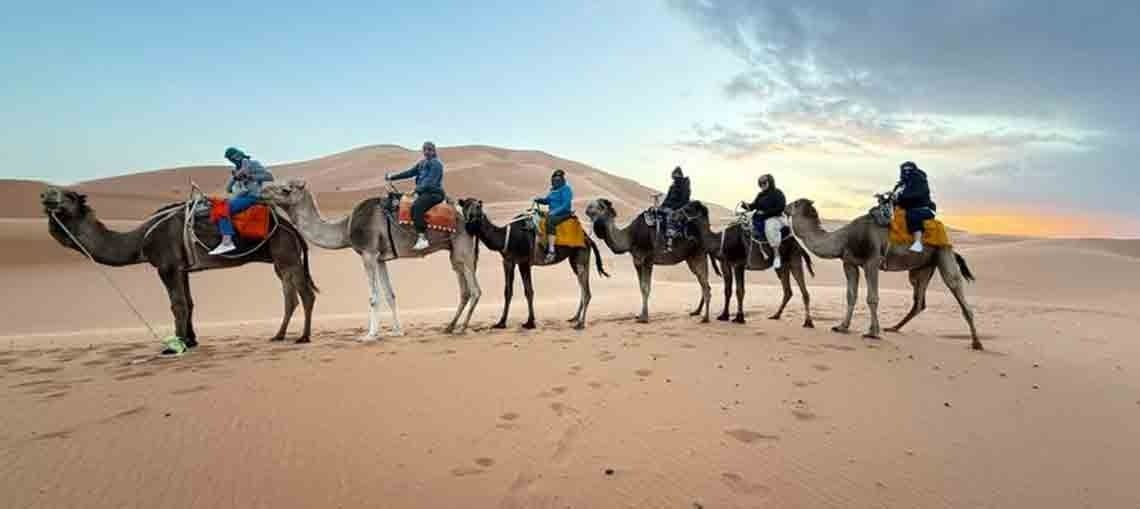 People riding camels across desert sand at sunset, with colorful saddles and a wide sky.