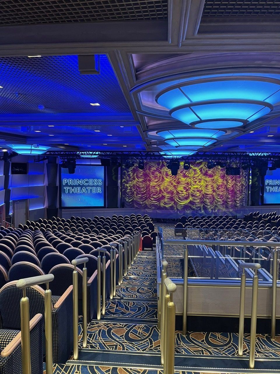 Empty auditorium with blue ceiling lights, rows of seats, and a stage with a colorful backdrop.