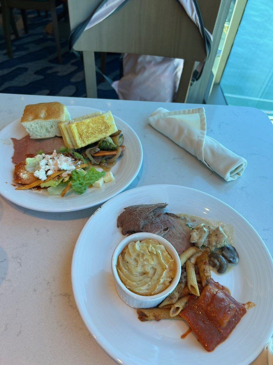 Two plates of cafeteria food on a table with a wrapped sandwich and a cup of soup
