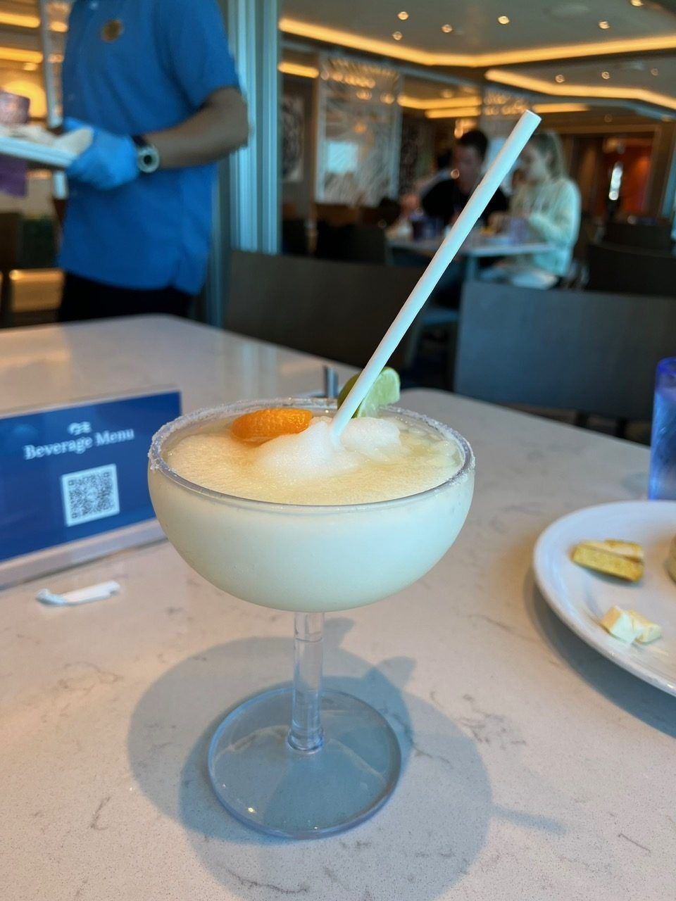 Foamy cocktail in a coupe glass with a straw on a restaurant table, next to a plate and QR code stand