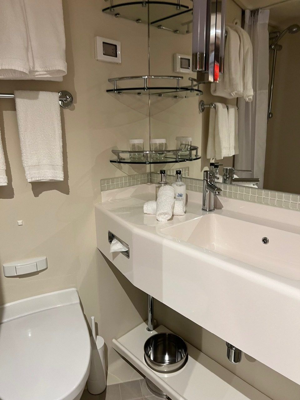 Small beige bathroom with white sink, toilet, mirror shelves, and hanging towels