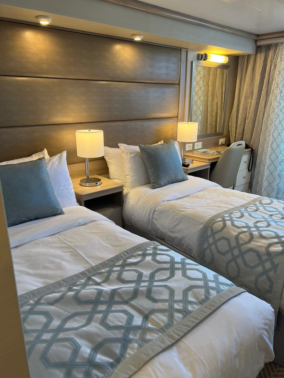 Twin beds in a beige hotel room with lamps, gray patterned bedding, and a small desk by the window.