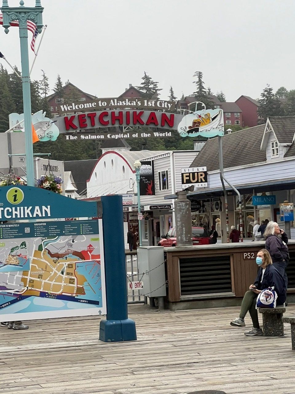 Tourist shop entrance in Ketchikan with colorful signs, map display, and people walking on a wooden boardwalk