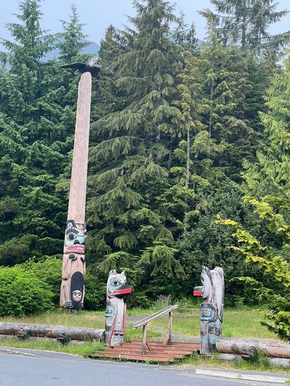 Totem pole and two bear statues in a park, framed by tall evergreen trees.